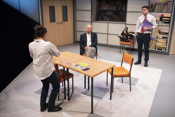 Three men are in a modern office; one is sitting at a table, another stands with arms folded, and the third stands holding folders. Bookcases, chairs, and a magazine on the table are visible in the background.