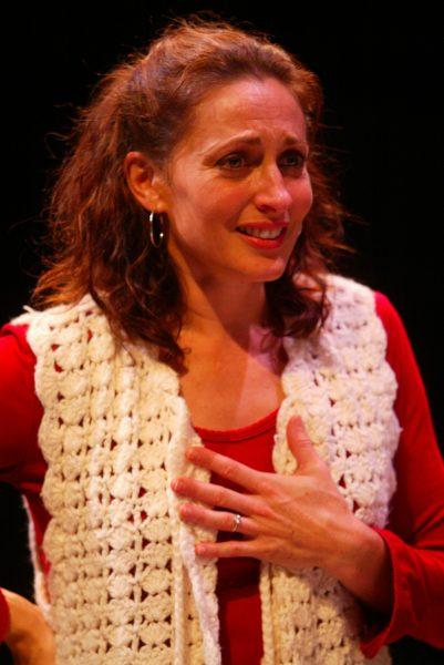 A woman with wavy brown hair wearing a white crocheted waistcoat and red top stands against a dark background, looking emotional with her hand on her chest and a slight, teary smile on her face.