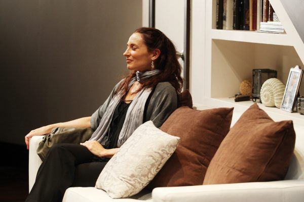A woman with long brown hair sits relaxed on a white sofa with brown and cream cushions, eyes closed, wearing a grey scarf and cardigan. Shelves with books and decor are seen behind her.
