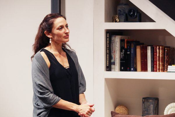 A woman with long brown hair, wearing a black top and grey cardigan, stands indoors near a white bookcase filled with books and decorative items. She is looking slightly to the side and smiling gently.