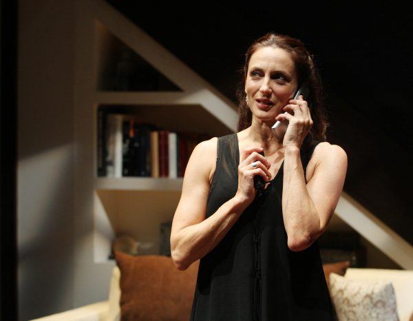 A woman in a sleeveless black dress stands indoors, holding a phone to her ear and smiling. Behind her are shelves with books and a white sofa with brown and white cushions.