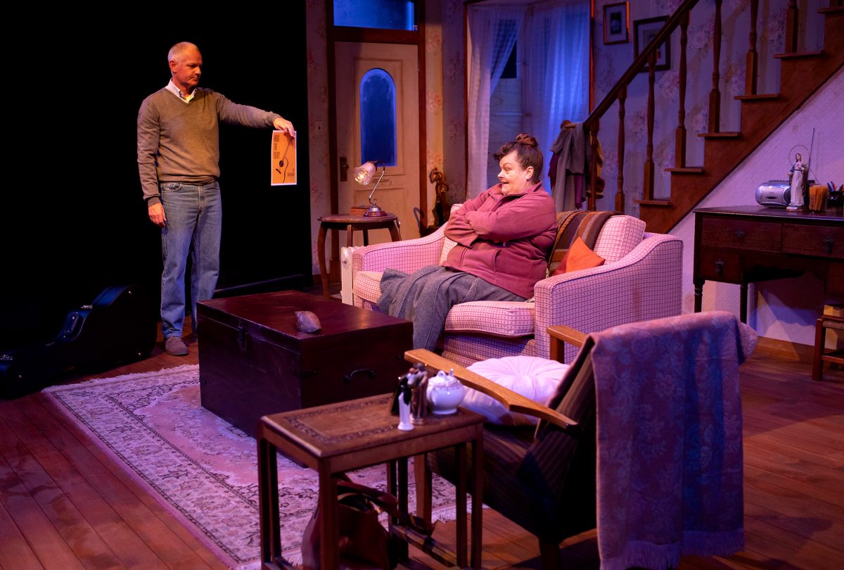 A man stands holding a framed picture while a woman sits on a sofa with her arms crossed in a cosy, warmly lit living room set with wooden floors, rugs, and a staircase in the background.