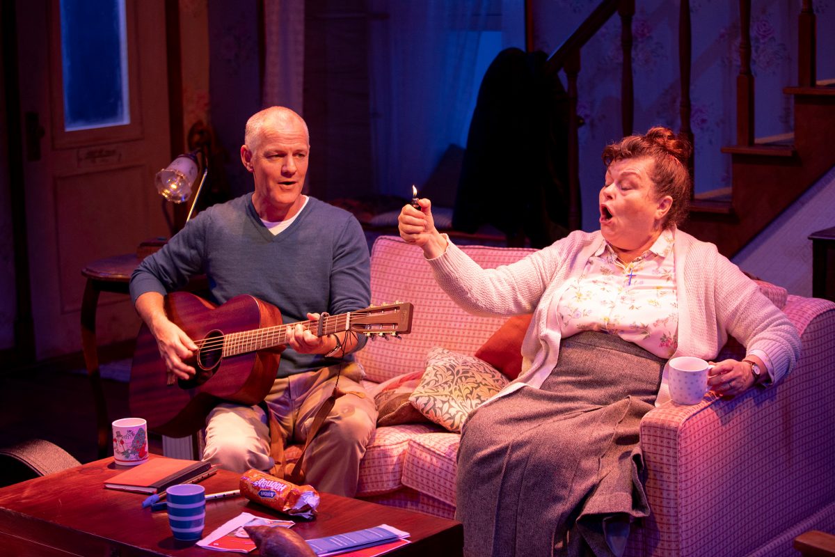 A man plays guitar on a pink sofa while an elderly woman beside him, holding a mug, excitedly lights a cigarette. The cosy living room has warm lighting, snacks, and papers on the coffee table.