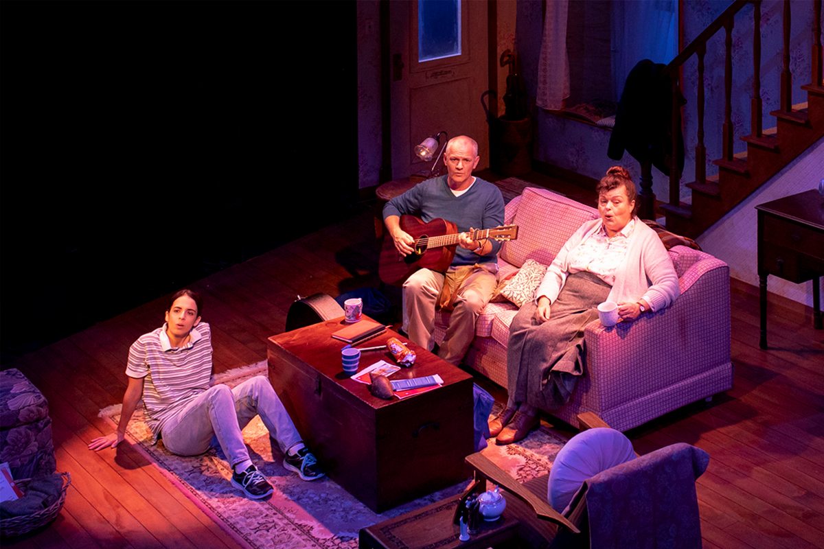 Three people sit in a warmly lit living room: a young man on the floor, an older man on the sofa playing a guitar, and a woman beside him holding a mug. They all look towards the front with serious expressions.