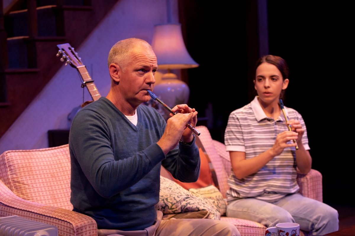 A man and a young woman sit on a pink sofa in a cosy living room, both playing tin whistles. The man wears a blue jumper and beige trousers; the woman wears a striped polo shirt. A guitar rests nearby and a lamp glows behind them.