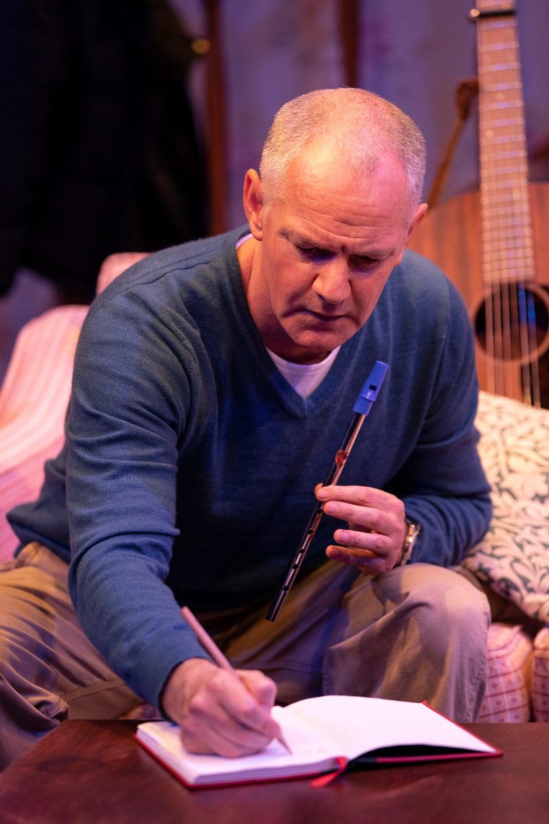 A middle-aged man with short grey hair sits on a sofa holding a tin whistle in one hand while writing in a notebook with the other. A guitar rests upright next to him in the background.