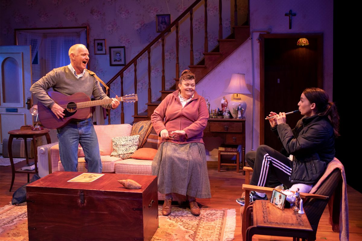Three people in a cosy living room: an older man playing guitar and singing, an older woman smiling and knitting, and a young woman in sportswear sitting and playing a recorder. The scene appears warm and lively.