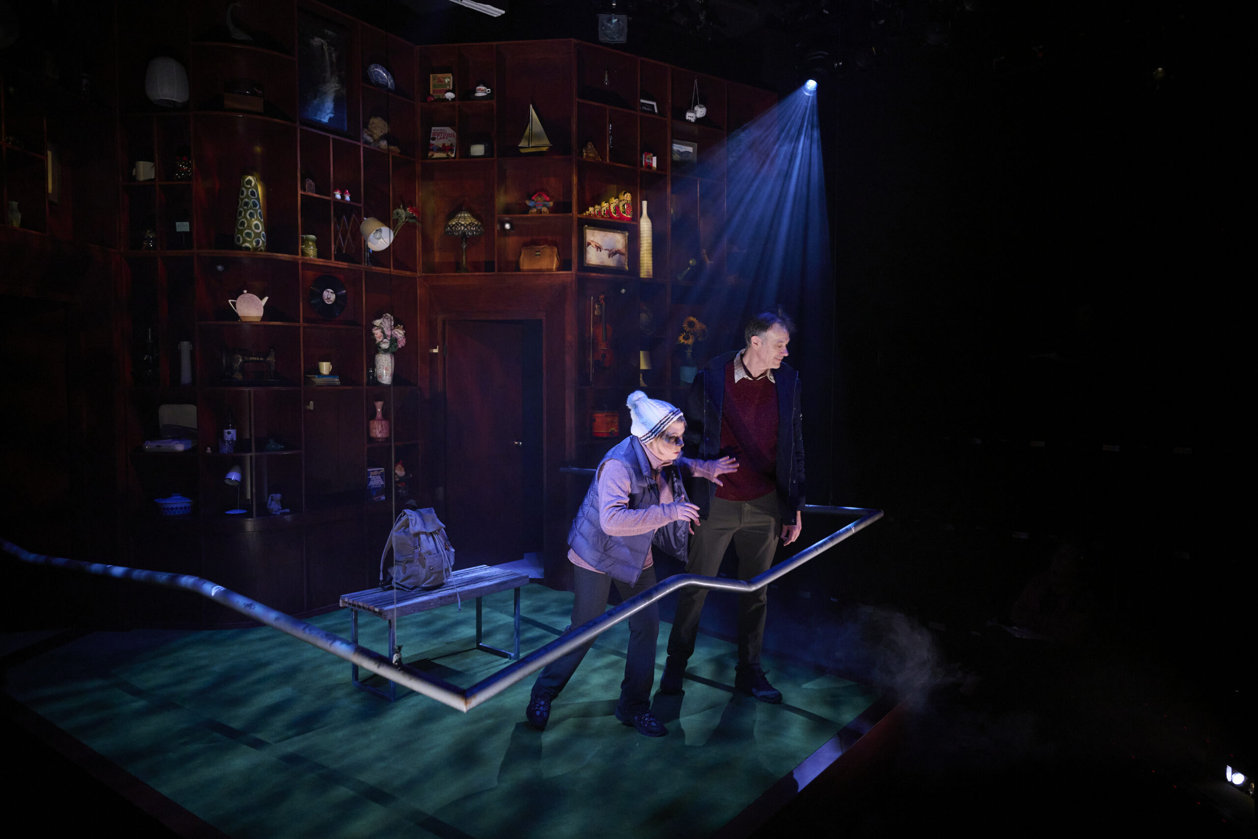 Two actors perform on a stage with dramatic lighting; one leans on a railing, wearing a white hat, whilst the other stands beside them. The backdrop is filled with shelves holding various objects and decorations.