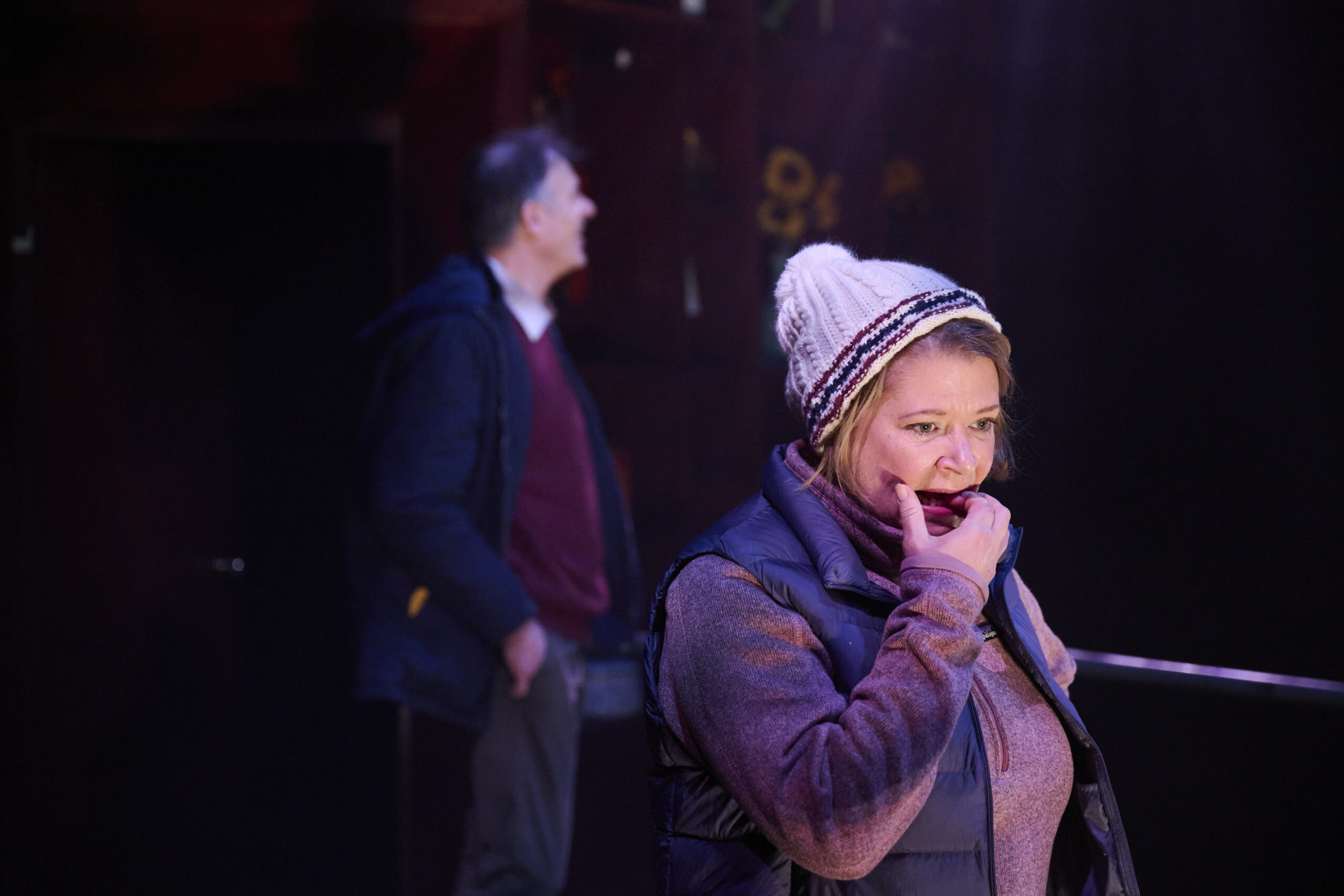 A woman in a knitted hat and waistcoat stands in the foreground, looking concerned with her hand near her mouth. A man in a jacket is blurred in the background, standing and looking off to the side. The lighting is dramatic.