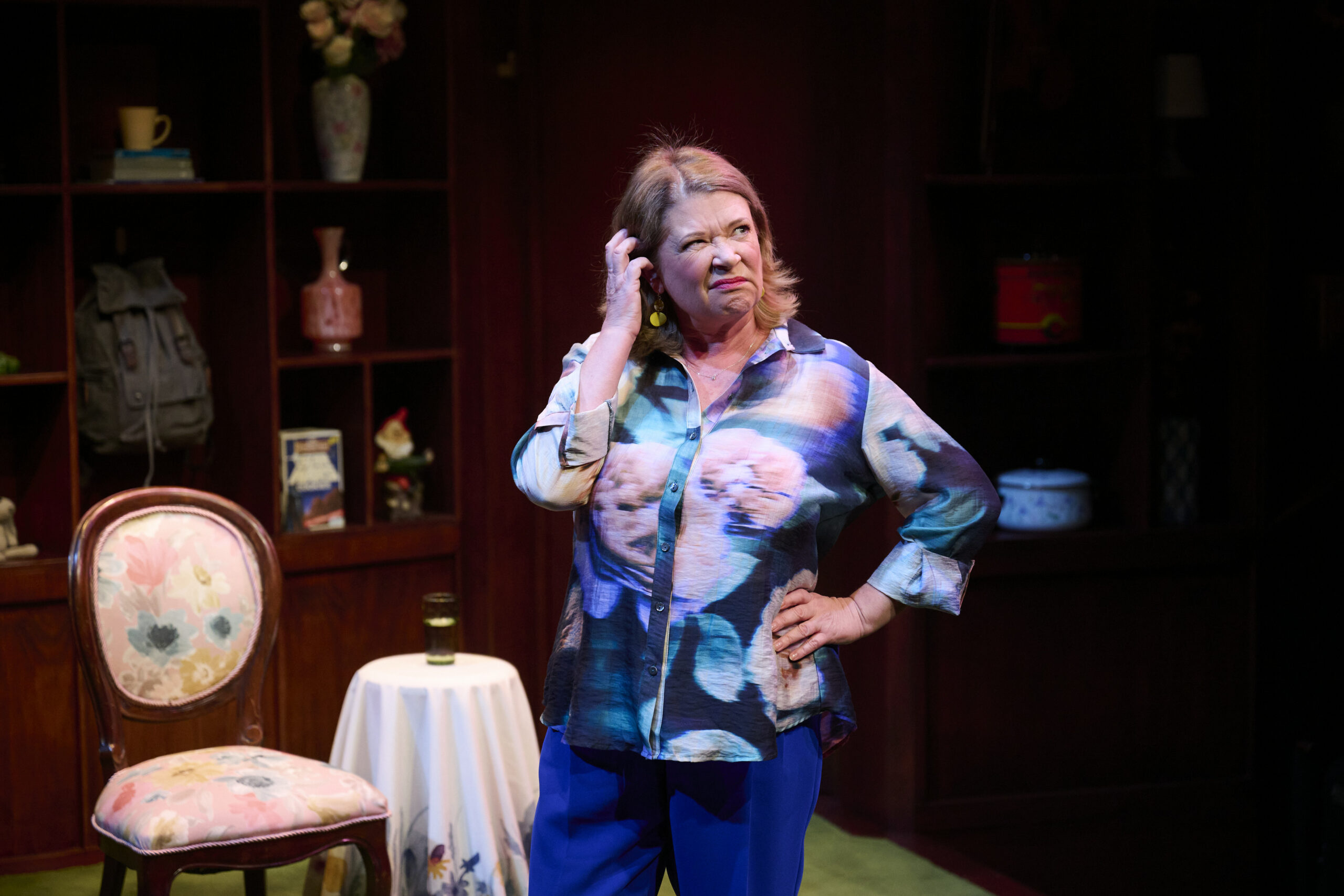 A woman in a colourful blouse stands with one hand on her hip and the other touching her head, looking puzzled. She is on stage in a cosy room with wooden shelves, a floral chair, and a small round table.
