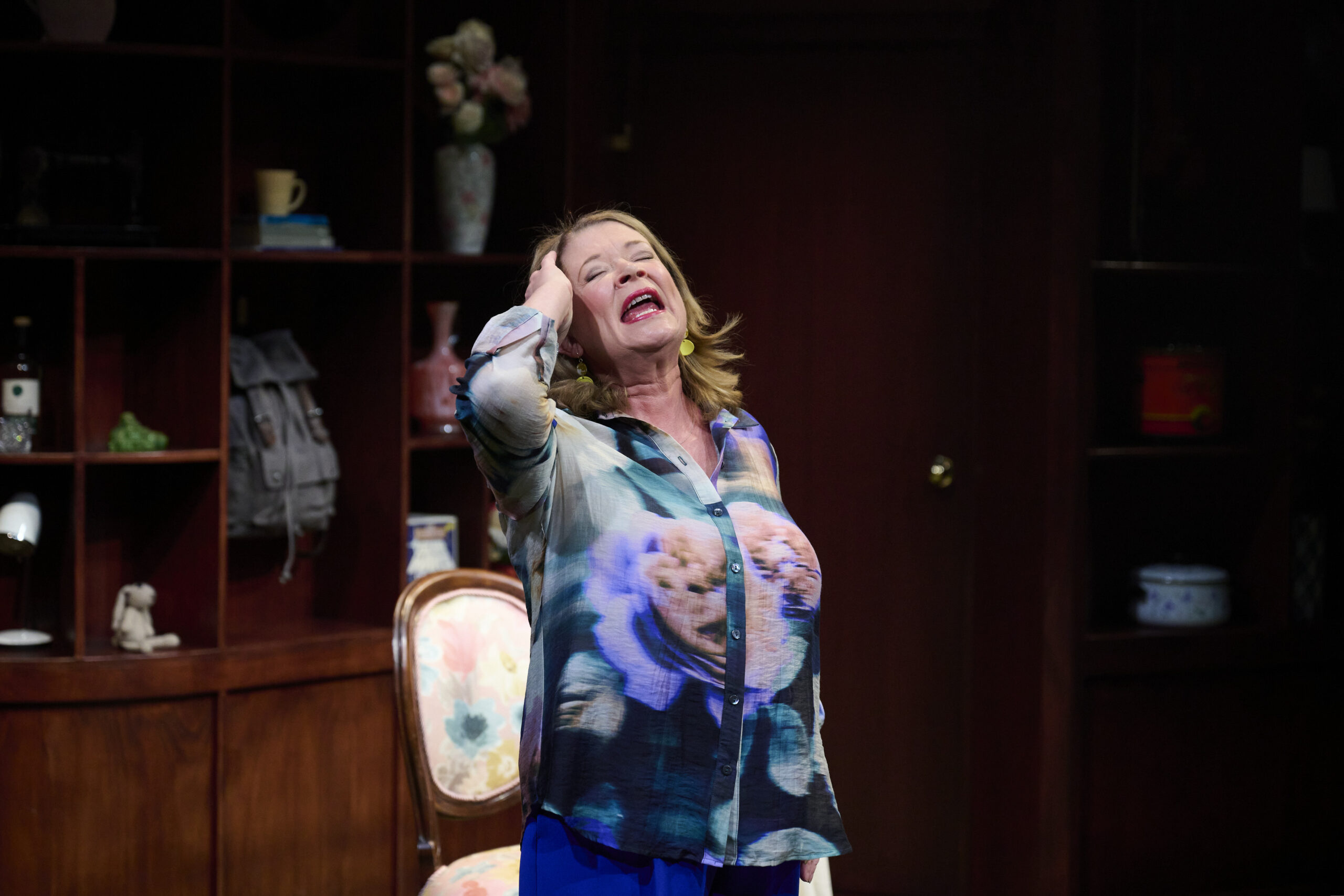 A woman stands in a room, smiling with her eyes closed and one hand on her head. She wears a colourful blouse with a large print and blue trousers. Behind her is a wooden shelf with various decorative items and a floral chair.