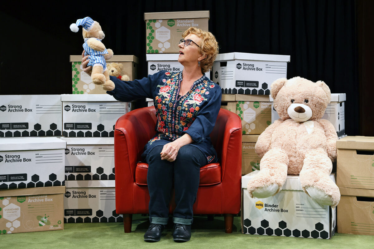 A woman with short curly hair sits on a red chair, holding up a small teddy bear in pyjamas. A large teddy bear sits beside her. Archive boxes are stacked in the background.