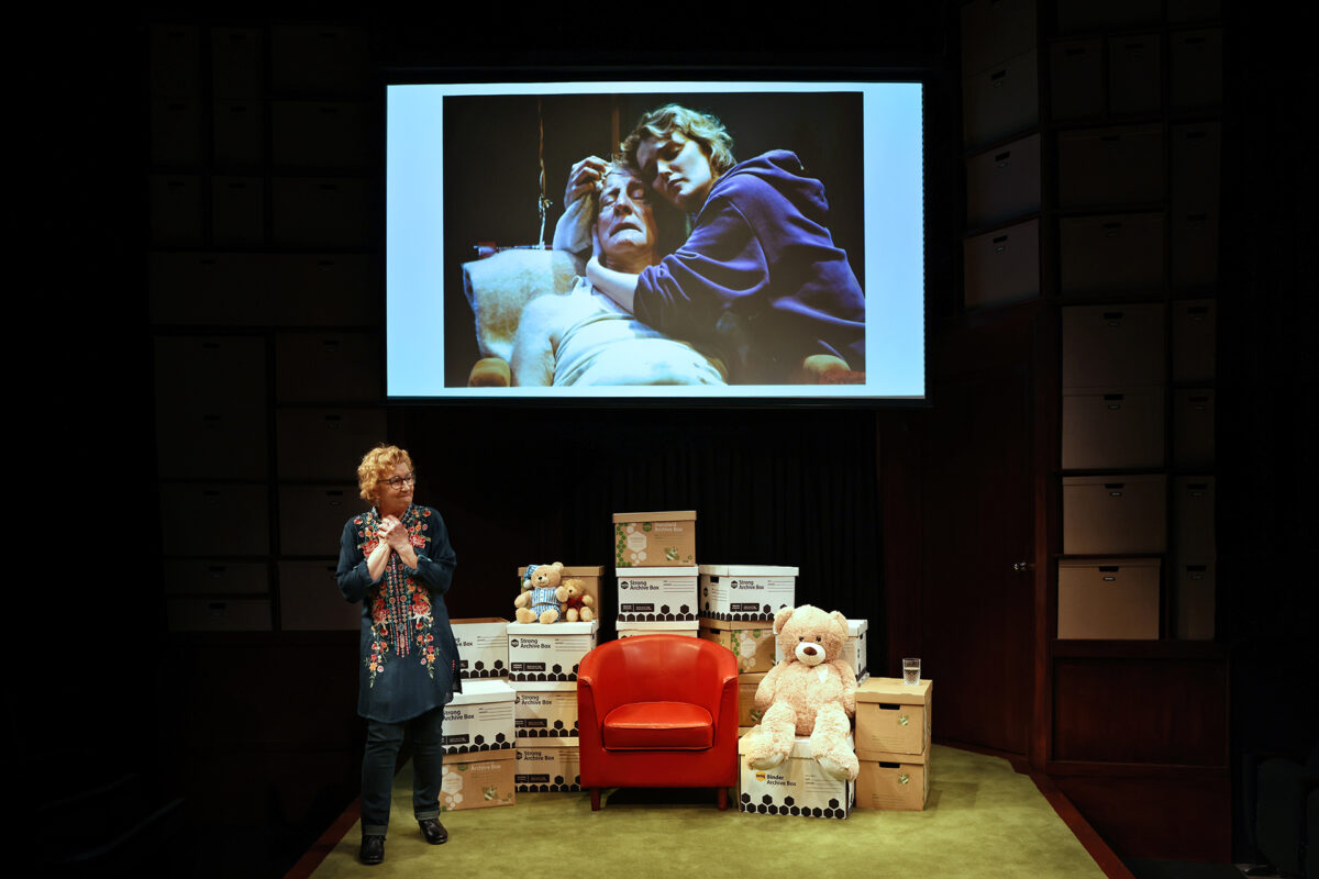 A woman stands on stage beside an orange chair, white boxes, and a large teddy bear, with a projected photo of a young person hugging an elderly woman in a hospital bed displayed behind her.
