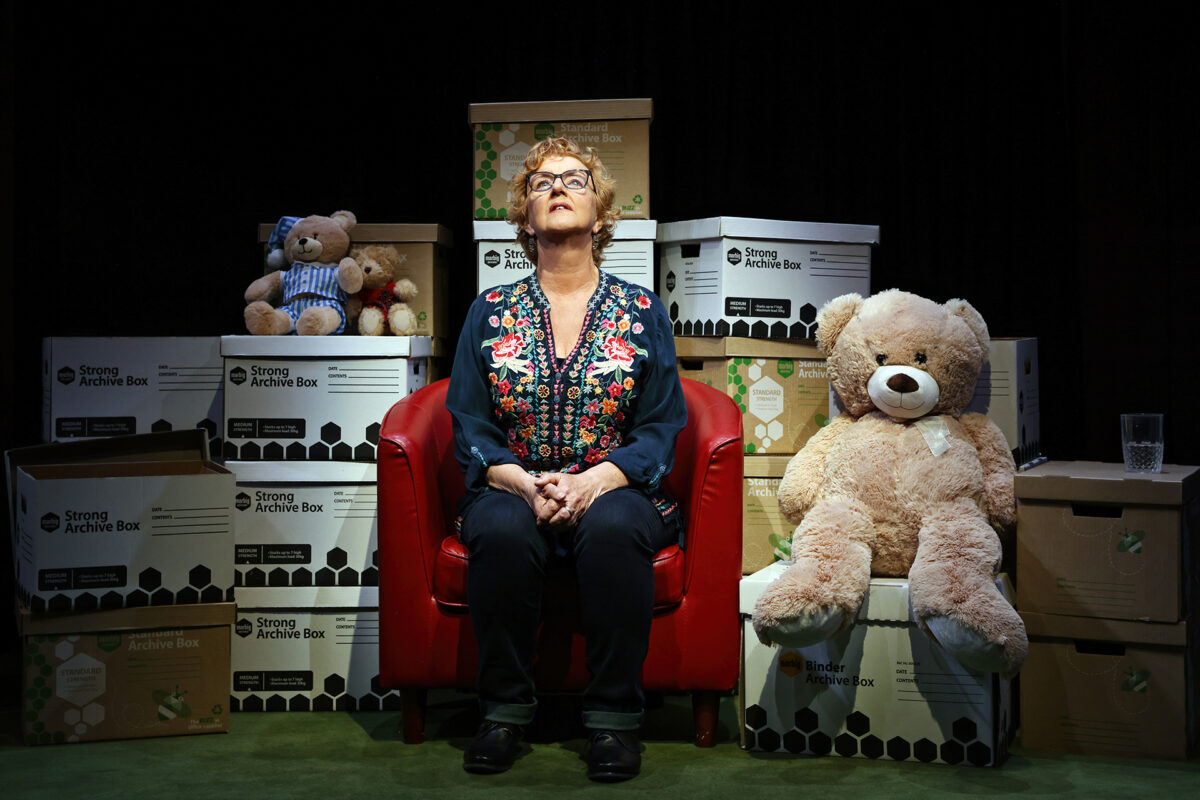 A woman sits on a red armchair, surrounded by archive boxes and two teddy bears. She looks upwards, wearing glasses and a floral blouse. The setting is indoors, with a glass of water on the armrest beside her.