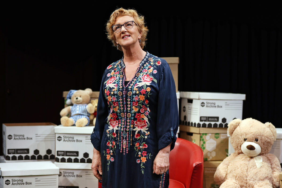 A woman in a dark floral dress stands smiling on stage, surrounded by archive boxes and two large teddy bears. A red chair is visible next to her, with a black background behind her.