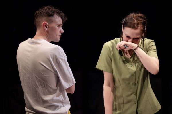 A young man in a white T-shirt looks at a young woman in a green shirt who is wiping tears from her eyes, appearing upset, against a dark background.