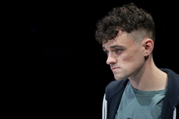 A young man with short curly hair and an earring looks serious and slightly tense, gazing to the right. He wears a green T-shirt and a dark hoodie, with a plain black background.