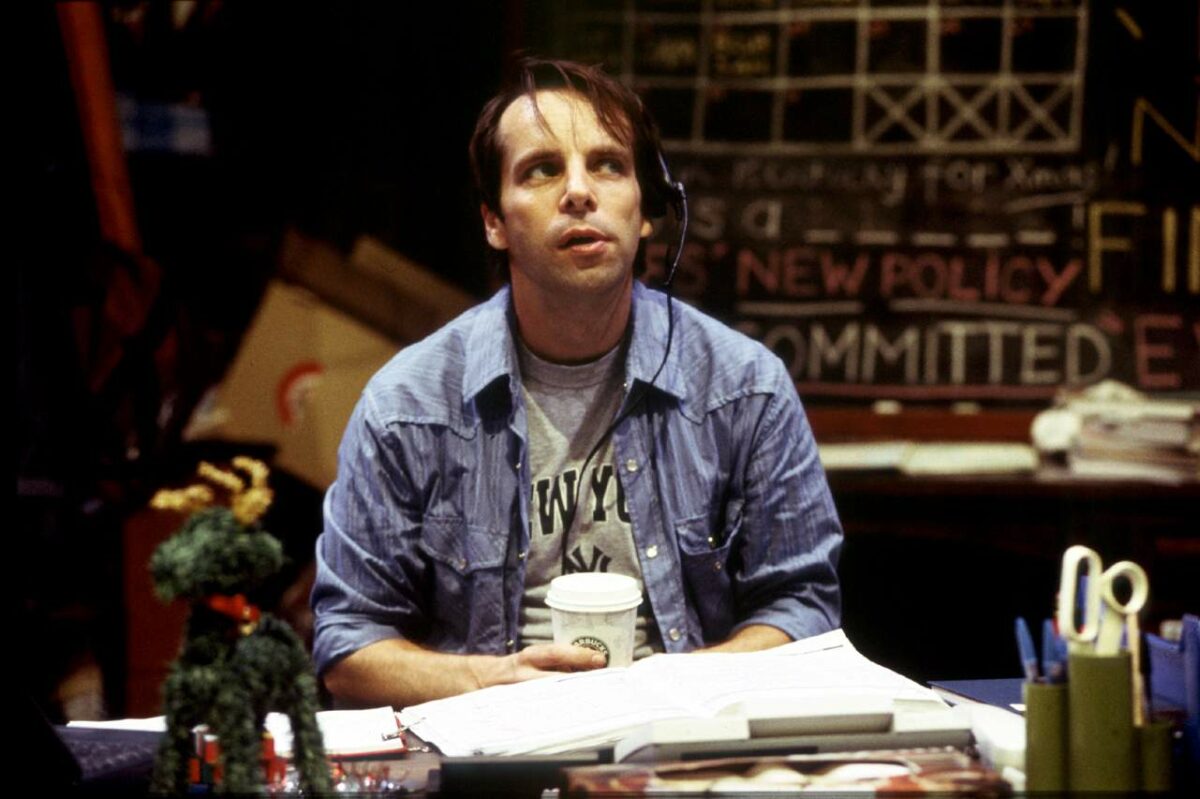 A man wearing a headset and holding a coffee cup sits at a cluttered desk with papers and office supplies. Behind him is a blackboard with colourful writing and diagrams. A small green puppet is visible on the desk.