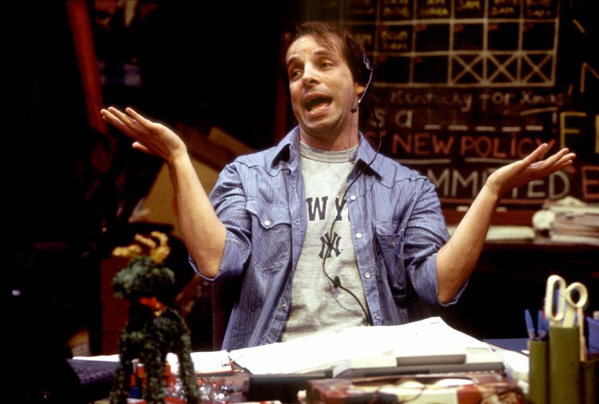 A man wearing a headset and a New York Yankees T-shirt shrugs his shoulders with his arms outstretched while sitting at a cluttered desk, with a large blackboard full of writing behind him.