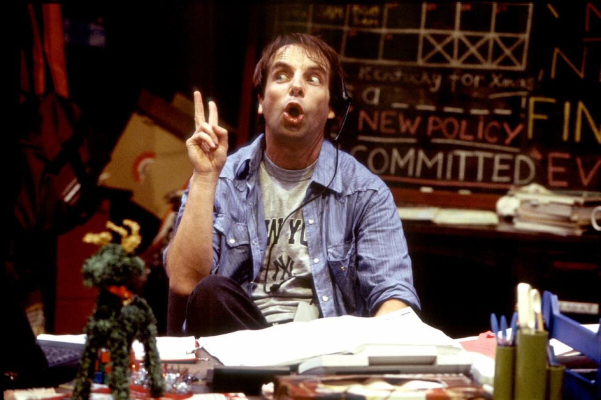 A man in a blue shirt sits at a cluttered desk, making a peace sign with his fingers and looking up with his mouth open. Behind him is a chalkboard with various colourful words and drawings.