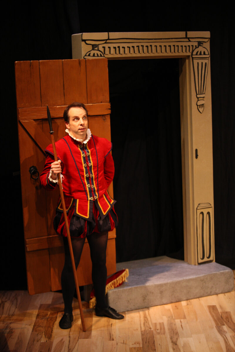 A man in a red Renaissance-style costume and holding a spear stands in a wooden doorway with a painted stone arch frame, performing on a stage with a wooden floor.