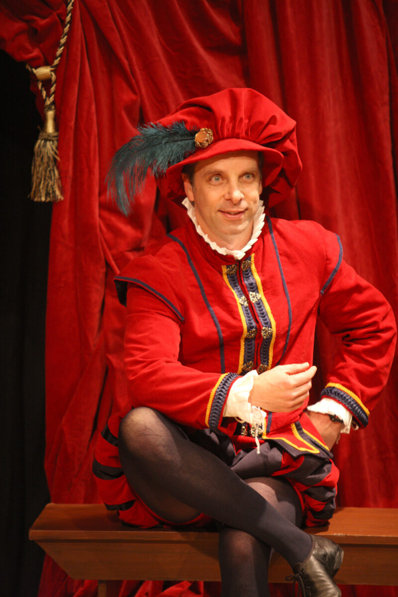 A person in an elaborate red Renaissance-style costume with a feathered hat sits on a bench, posing confidently in front of matching red curtains.