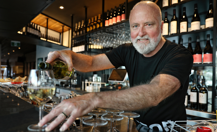 A smiling barman with a beard pours white wine into a glass at a bar, surrounded by empty glasses and shelves filled with bottles of wine in the background.