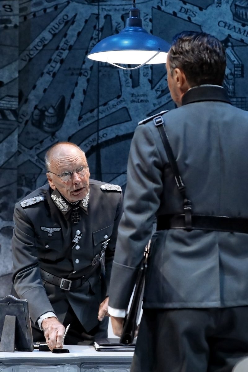 An older man in a military uniform leans on a desk, speaking intensely to another man in uniform who stands with his back to the camera. A hanging lamp illuminates the scene, and a detailed map covers the wall behind them.