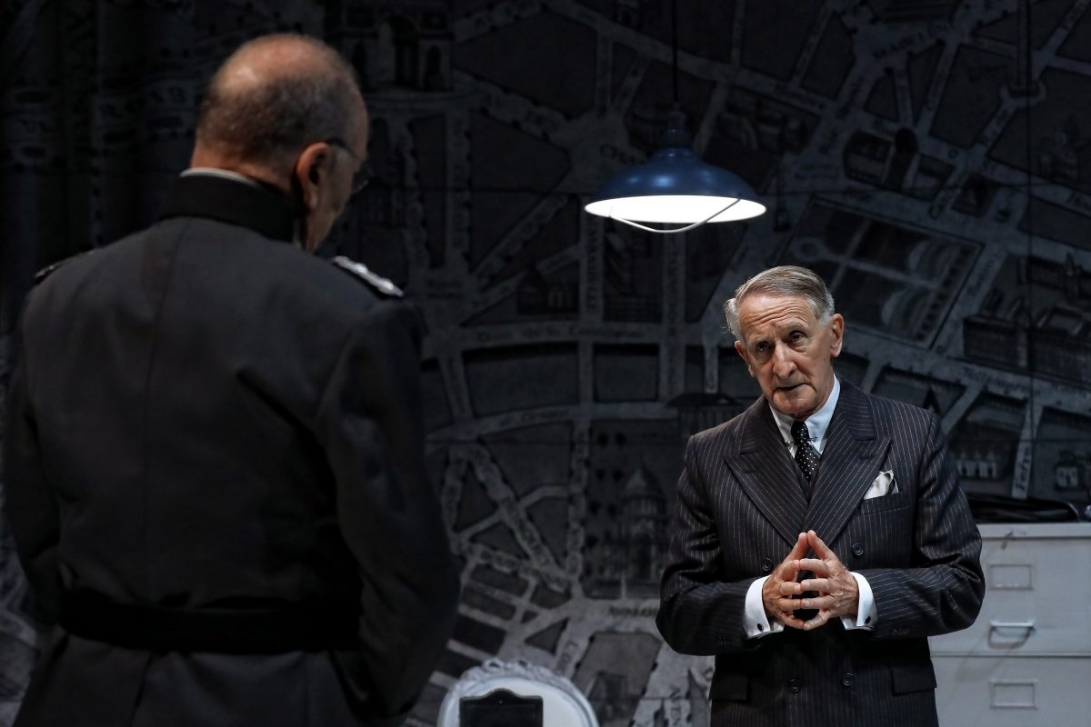 Two older men in suits face each other under a hanging light. One man, standing with hands clasped, appears to be speaking seriously, whilst the other, seen from behind, listens. The background features a large, dark map.