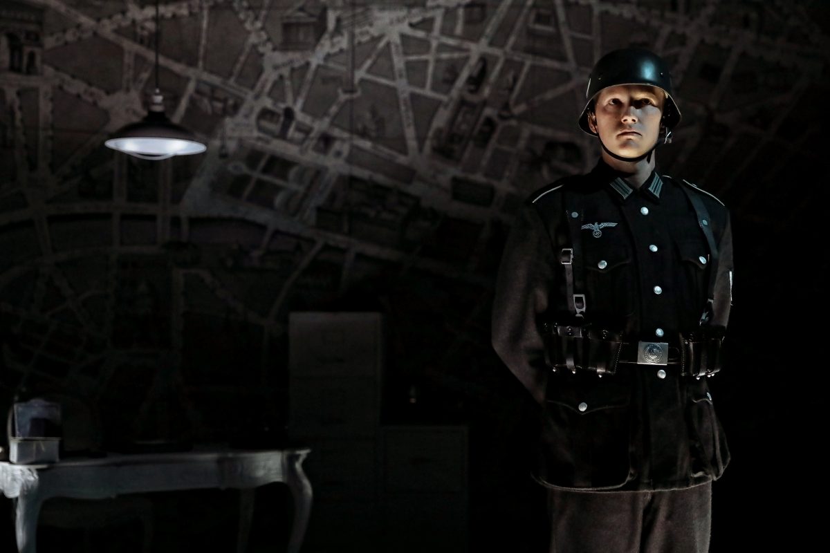 A person in a dark military uniform and helmet stands to attention in a dimly lit room with a wall covered in maps, next to a desk with a lamp and some papers.