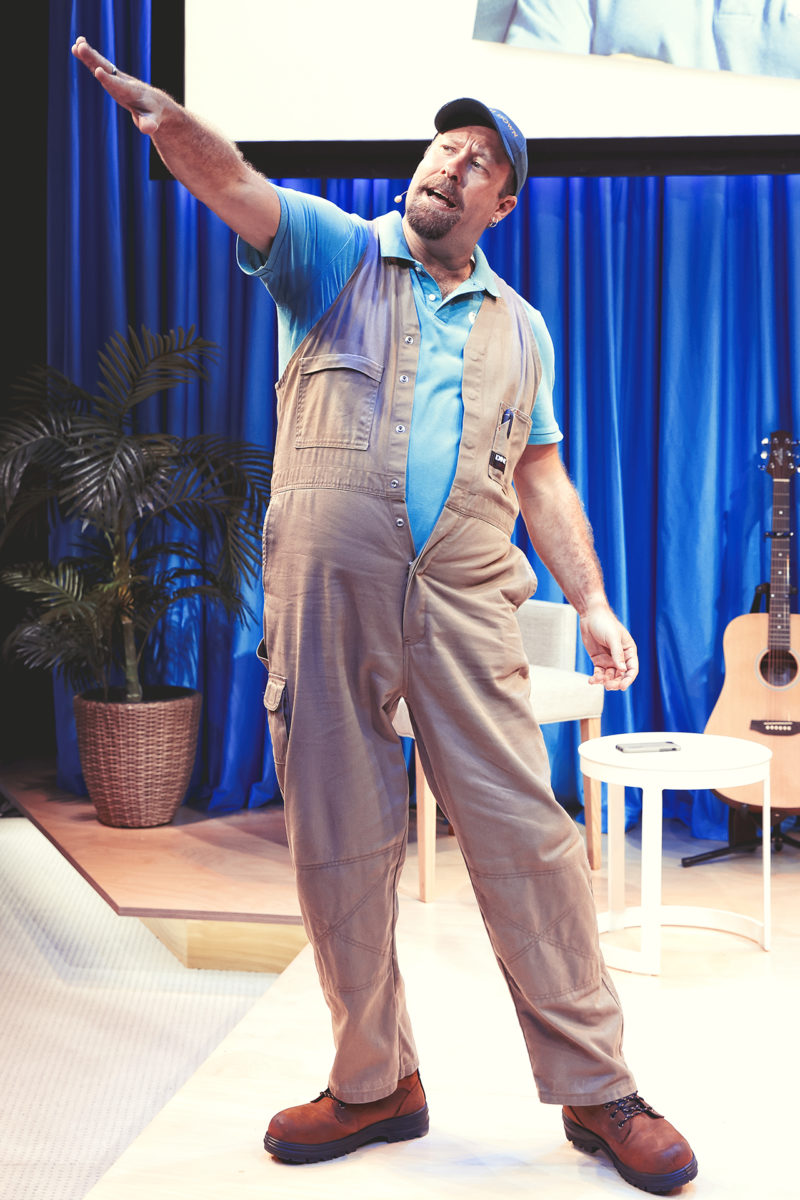 A man in brown overalls, blue shirt, and cap stands on a stage with his arm raised, gesturing energetically. Behind him are blue curtains, a potted plant, a white chair, a small table, and an acoustic guitar.