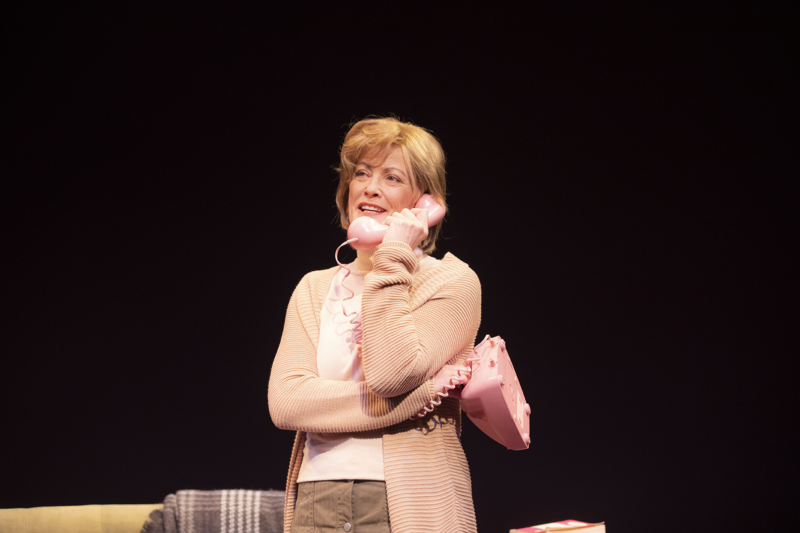 A woman with short blonde hair, wearing a light pink cardigan and white shirt, smiles whilst holding a vintage pink rotary telephone to her ear on a dark stage.