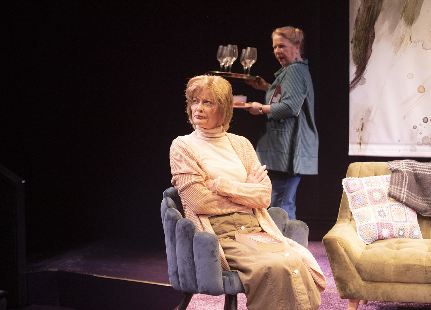 A woman with short blonde hair sits on a blue chair with her arms folded, looking serious. Another woman in the background carries a tray with empty wine glasses. They are on a stage with cosy furniture.
