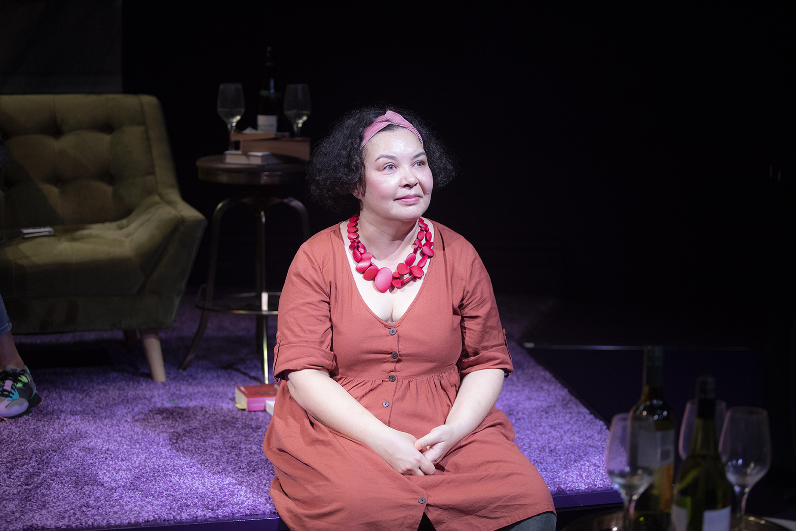 A woman with curly hair and a pink headband sits on a purple carpet, wearing an orange dress and chunky pink necklace. She is lit by stage lights, with wine glasses, bottles, and an armchair in the background.