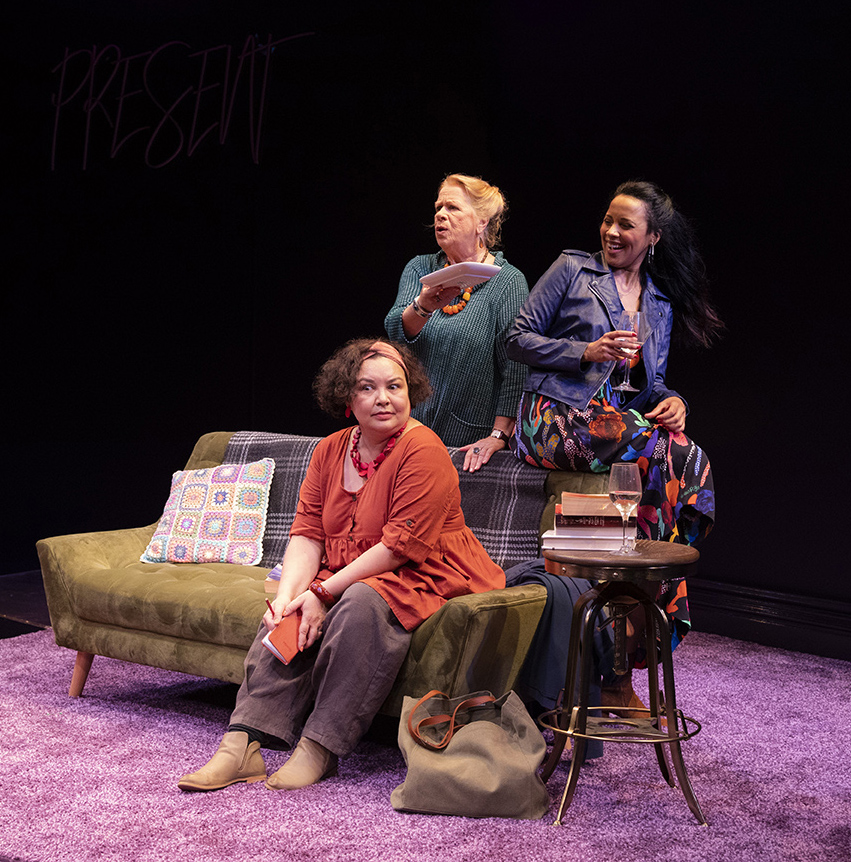 Three women are on a stage set. One sits on a green sofa looking thoughtful, another stands behind the sofa holding papers and looking surprised, whilst the third stands smiling with a drink. The scene is warmly lit with a purple carpet.
