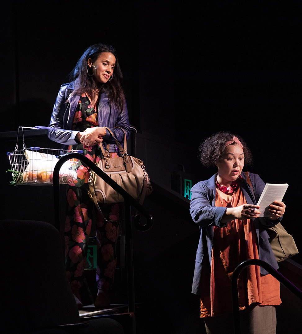 Two women on stage: one stands holding a book and reading aloud, wearing a red scarf and blue jacket; the other, in a floral dress and purple jacket, stands above her with a large handbag and a shopping basket, looking down and smiling.