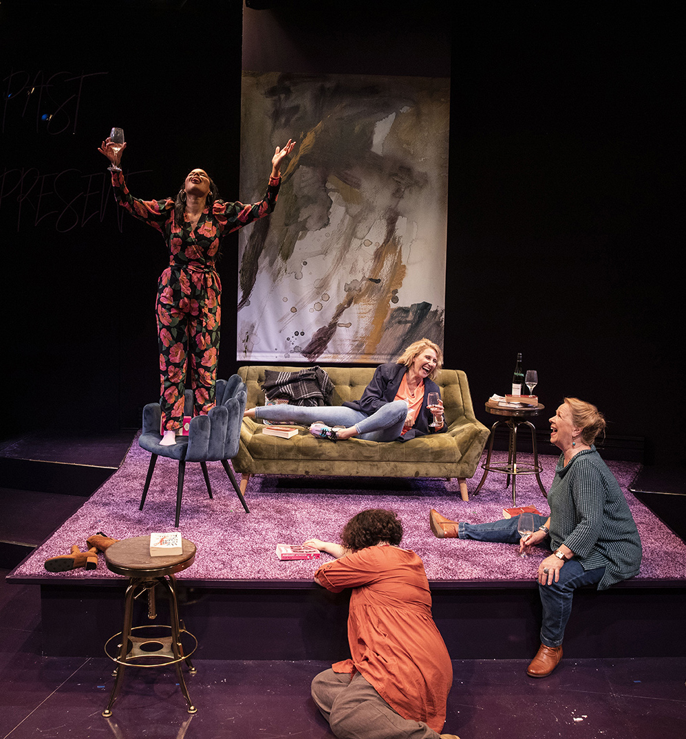 Four women are on a stage set resembling a lounge. One woman stands on a chair with arms raised, while the others sit or kneel on the floor, laughing and holding wine glasses. The scene is lively and colourful.