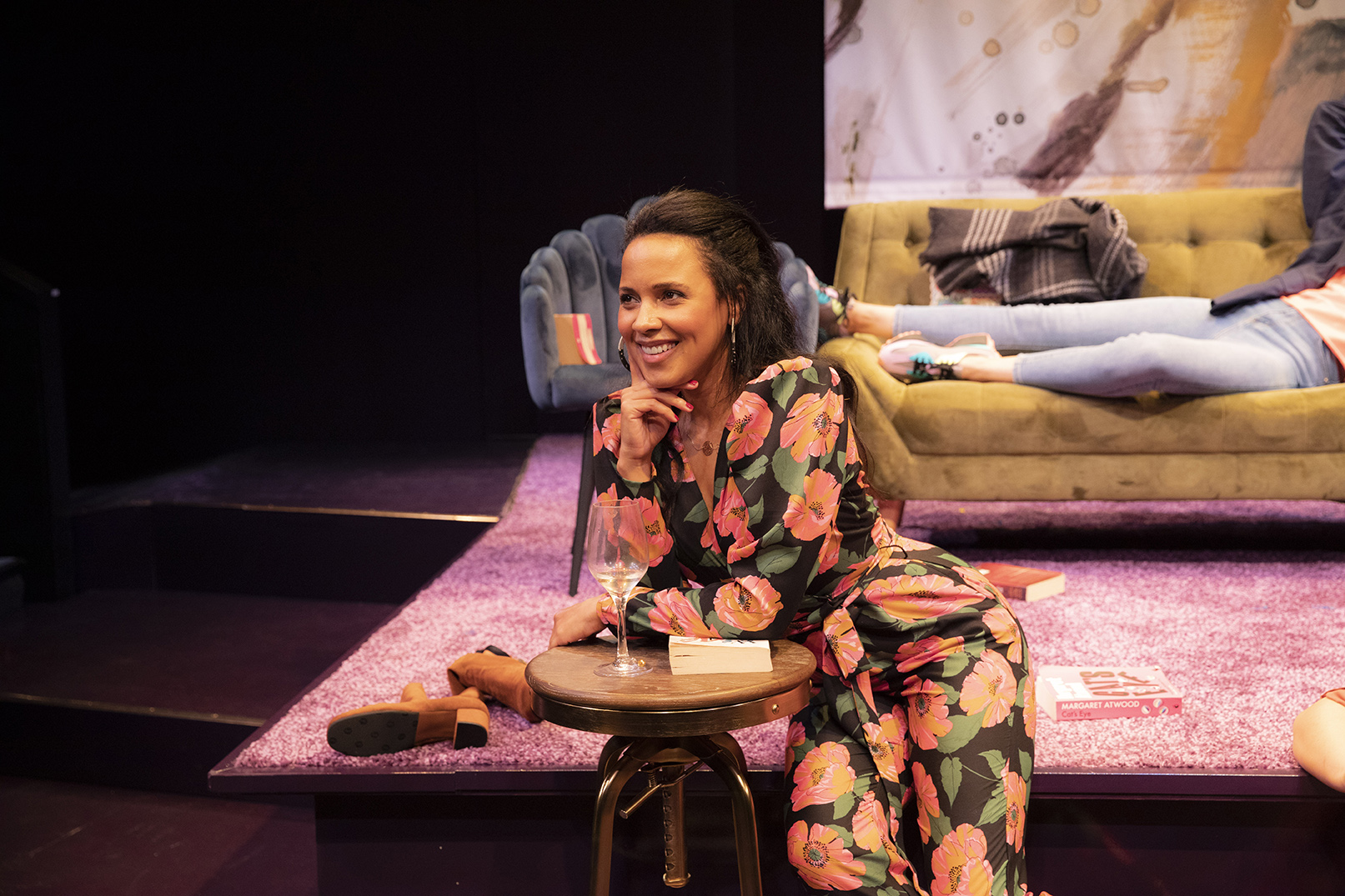 A woman in a floral suit sits smiling by a small round table with a wine glass, on a purple carpet. Behind her, another person lounges on a beige sofa, with a casual, cosy stage set in the background.