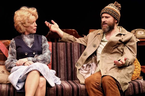 A woman and a bearded man sit on a striped sofa in conversation; the woman wears a dress and holds a blanket, while the man gestures expressively, wearing a knitted hat and trench coat.
