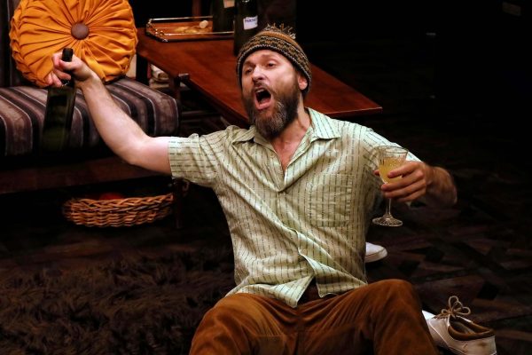A bearded man wearing a knitted hat sits on the floor, holding a wine bottle in one hand and a glass in the other, with his mouth open as if shouting or singing. Shoes and a furry rug are nearby.