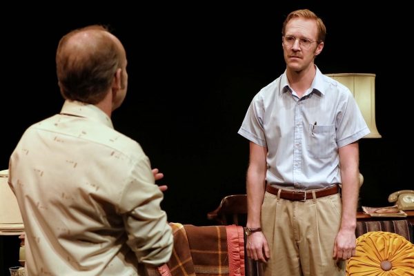 Two men are having a serious conversation in a dimly lit room. One man faces away from the camera, while the other, wearing glasses and a tucked-in shirt, looks at him with a concerned expression.