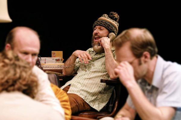 A bearded man in a winter hat speaks on a beige rotary phone, gesturing with his hand, while three other men sit nearby, two with heads lowered, all in a dimly lit room with a dark background.