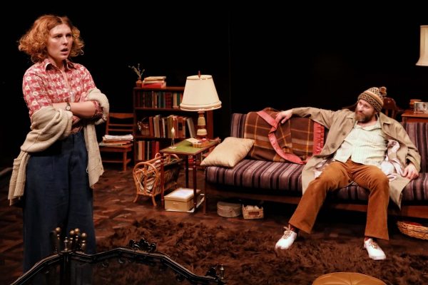 A woman with curly hair stands with arms crossed and a concerned expression, while a bearded man in a beanie and casual clothes reclines on a striped sofa in a cosy, cluttered living room set on stage.