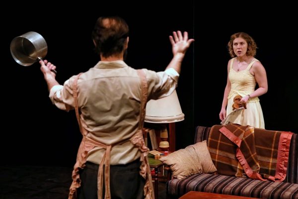 A man with his back to the camera lifts a saucepan while facing a surprised woman in a yellow dress, standing near a sofa covered in patterned blankets in a dimly lit room.