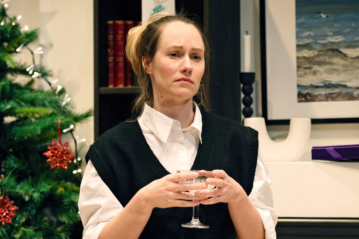 A woman with a serious expression holds a glass in both hands. She wears a white shirt with a black waistcoat. A decorated Christmas tree and bookcase are visible in the background.