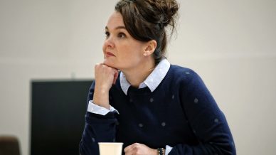 A woman with brown hair in a loose bun sits at a table, resting her chin on her hand and looking thoughtfully to the side. Papers and a paper cup are on the table in front of her. She wears a navy jumper over a collared shirt.