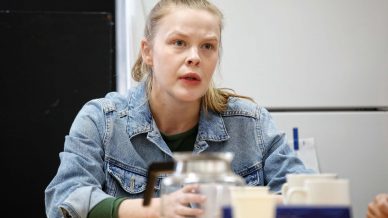 A woman with blonde hair in a ponytail, wearing a denim jacket, sits at a table with a glass coffee pot, cups, and papers. She appears to be speaking or listening intently indoors.