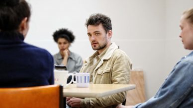 Four people sit around a table in a casual meeting. One man in a beige jacket looks attentively forward, while the others appear to listen or reflect. Mugs and a chair are visible, creating a relaxed atmosphere.