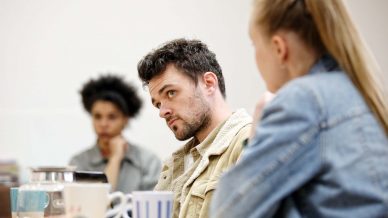 Three people sit at a table with coffee mugs, engaged in conversation. The man in the centre looks attentively to his right, while a woman with a ponytail sits beside him, and another woman is blurred in the background.