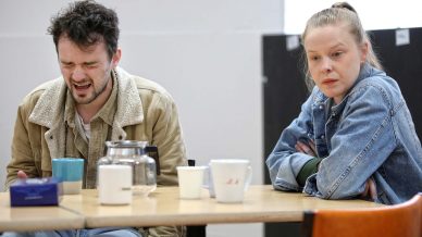 A man with a pained expression sits at a table next to a woman with folded arms who looks concerned. Several cups and a glass jug are on the table in front of them.
